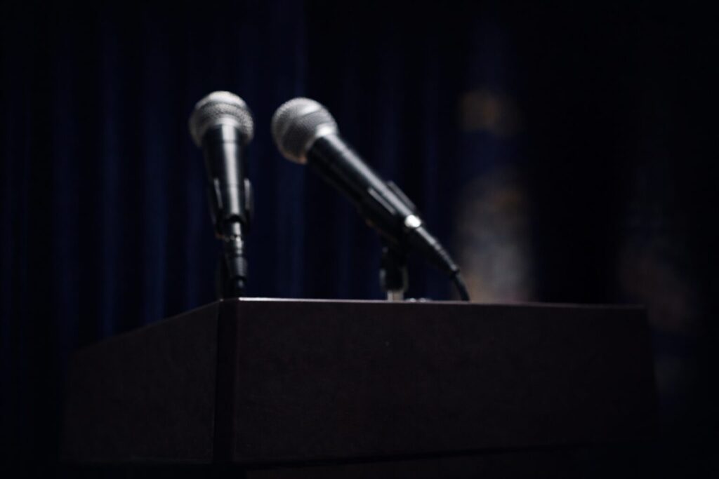 Out-of-focus podium microphones in front of a dark backdrop, suggesting public messaging that feels performative rather than credible.